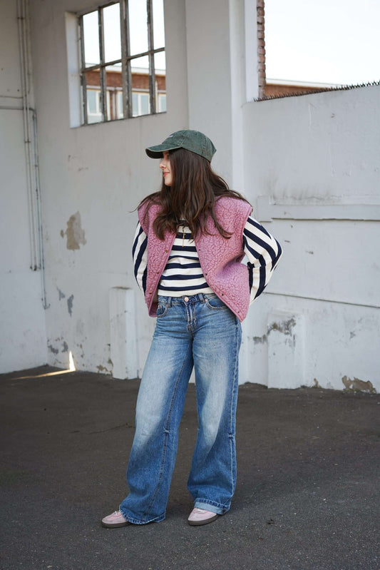 Vrouw in roze wollen bodywarmer, gestreepte trui en wijde jeans staat in een loods